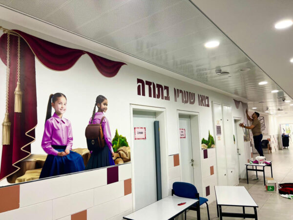 קיר מעוצב בחיתוכים צורניים עם הכיתוב בואו שעריו לתודה ודמויות של ילדות בצדדים , ווילונות עיצוב שמוסיף המון לאווירת בית הספר